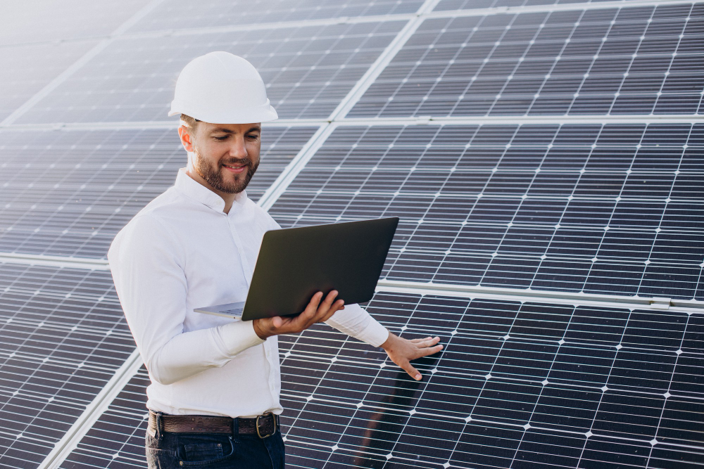 um homem com camisa social e capacete de obra segurando um tablet enquanto a outra mão está apoiada sobre o painel de energia solar